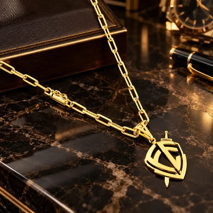 Vista superior do colar banhado a ouro 18k com corrente Cartier, fecho canhão e pingente vazado em formato de escudo de fé atravessado por espada sobre mármore escuro.