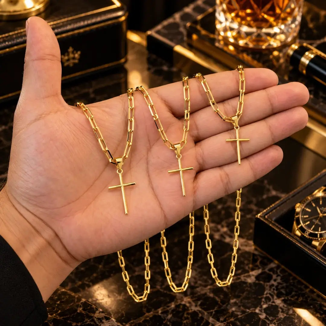 Mão masculina segurando três colares banhados a ouro 18k com corrente Cartier e pingente cruz palito para comparação de tamanho e proporções.