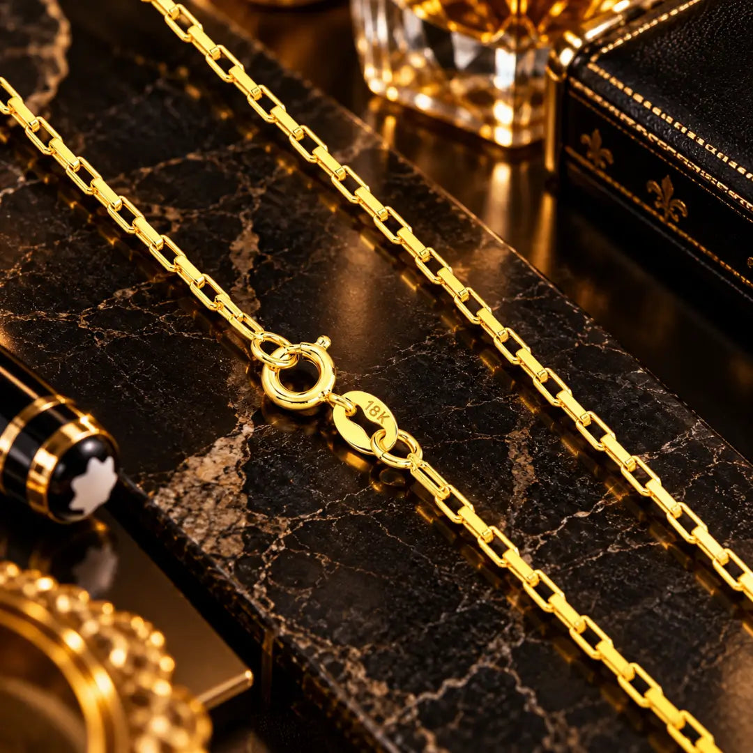 Detalhe em aproximação do fecho boia banhado a ouro 18k com gravação de autenticidade 18K e dos elos retangulares estilo Cartier tijolinho sobre bancada de mármore escura.