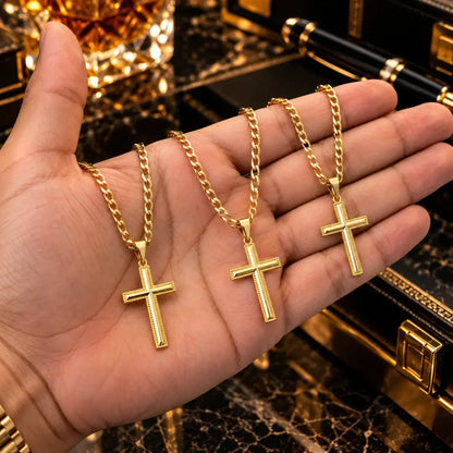 Mão masculina segurando três colares banhados a ouro 18k com corrente grumet e pingente de cruz detalhada para comparação de proporção e tamanho.