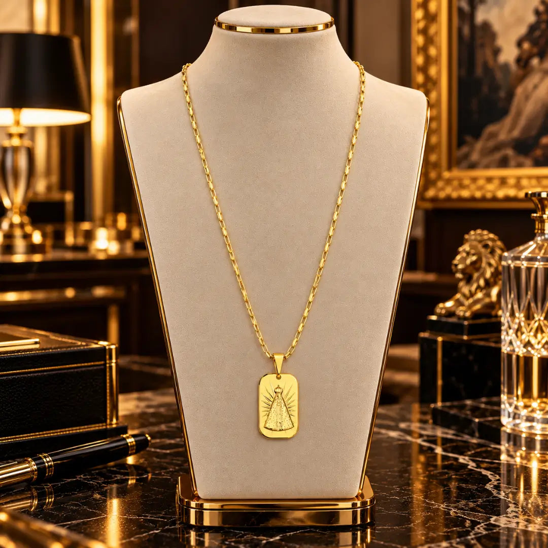 Colar masculino banhado a ouro 18k com corrente tijolinho e pingente medalha de Nossa Senhora Aparecida em exibição num busto expositor bege.