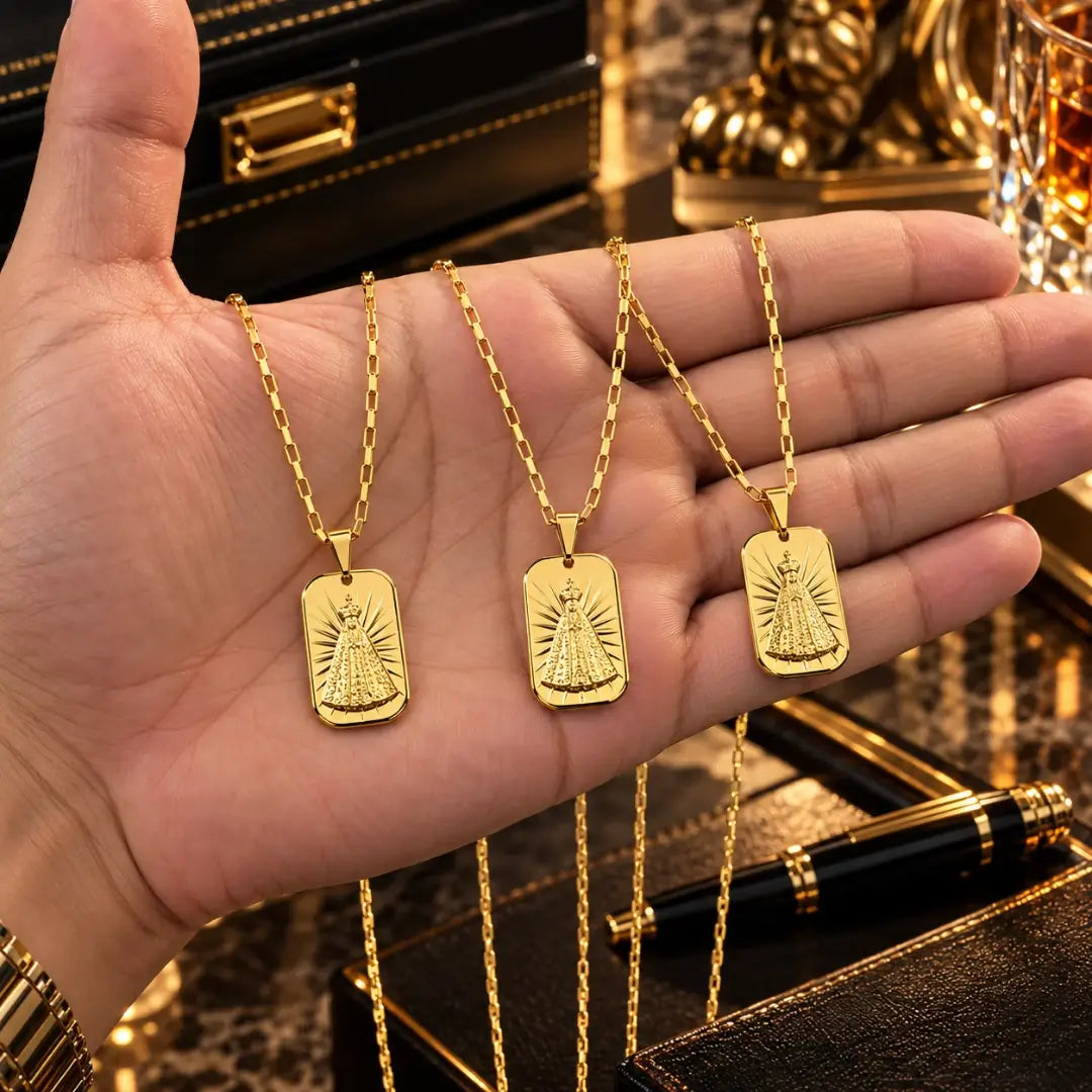 Mão masculina segurando três colares banhados a ouro 18k com corrente tijolinho e pingente de Nossa Senhora Aparecida para visualização de proporção.