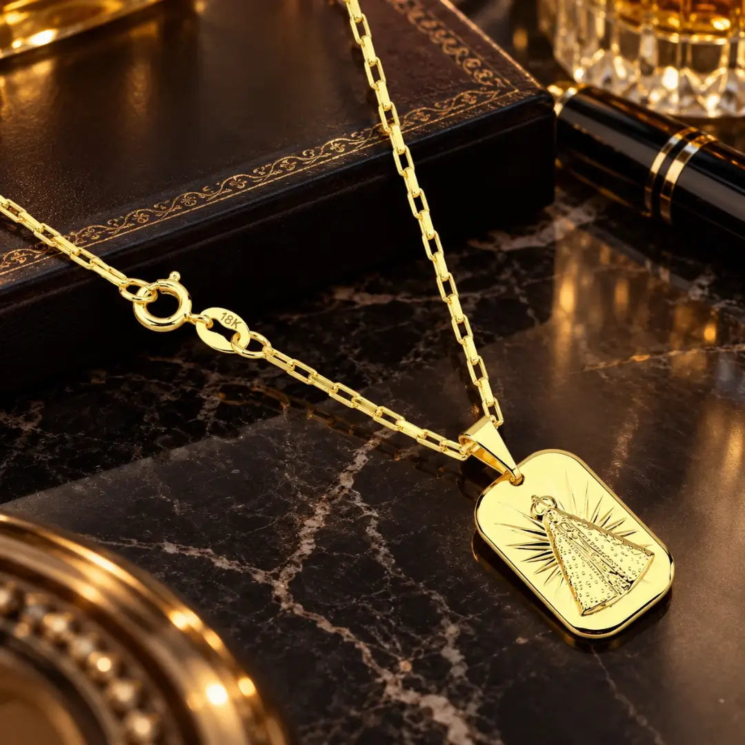 Visão geral do colar de corrente tijolinho e pingente de Nossa Senhora banhados a ouro 18k posicionados sobre bancada de mármore luxuosa.